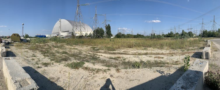 Aperçu d’un site semi-abandonné, avec un peu d’herbe et les structures de la centrale dans le fond, toujours reliées à des pylônes électriques